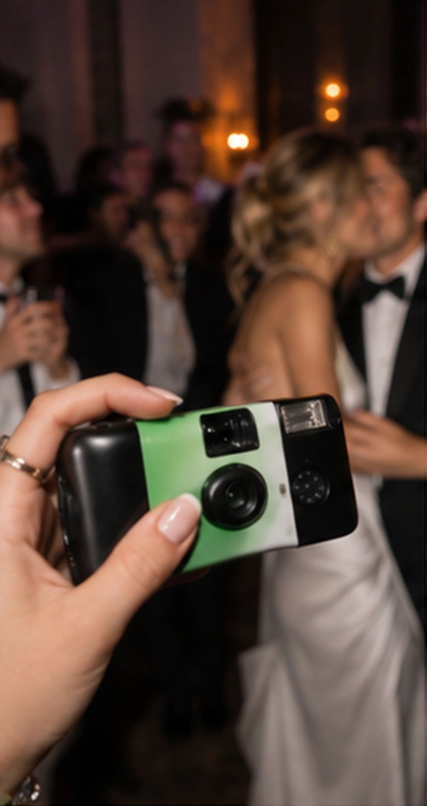 A hand holding a green disposable camera at a late-night wedding, a couple kissing behind