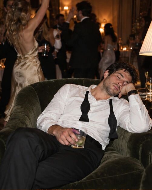 A guest asleep in a velvet armchair, bow tie undone, glass still in hand
