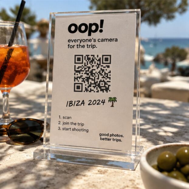 An acrylic stand on a beach-bar table with an oop card and a QR code that opens oop.camera/holidays, beside an Aperol spritz and olives