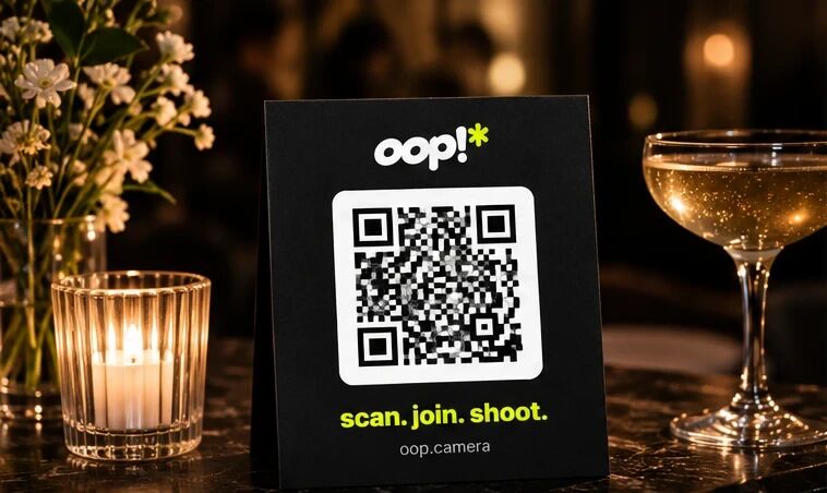 A black oop tent card with a QR code and “scan. join. shoot.” beside a candle and a coupe of champagne